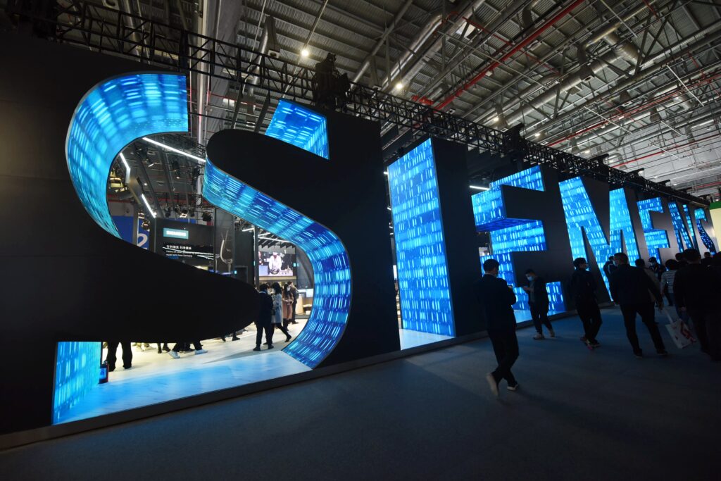Logo von Siemens künstlerisch dargestellt indem die Buchstaben ausgeschnitten sind und mit hellen und dunkelblauen Grafiken gestaltet sind. Über dem riesigen Logo ist Stahl und Beleuchtung. Davor spazieren viele Menschen